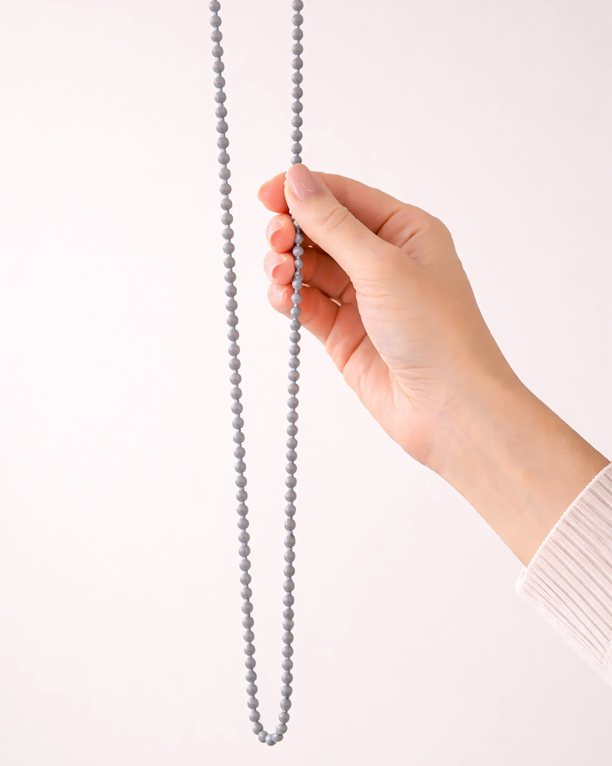 Corrente de acionamento manual cinza para persiana sob medida, em detalhe, sendo manuseada com uma mão em fundo neutro