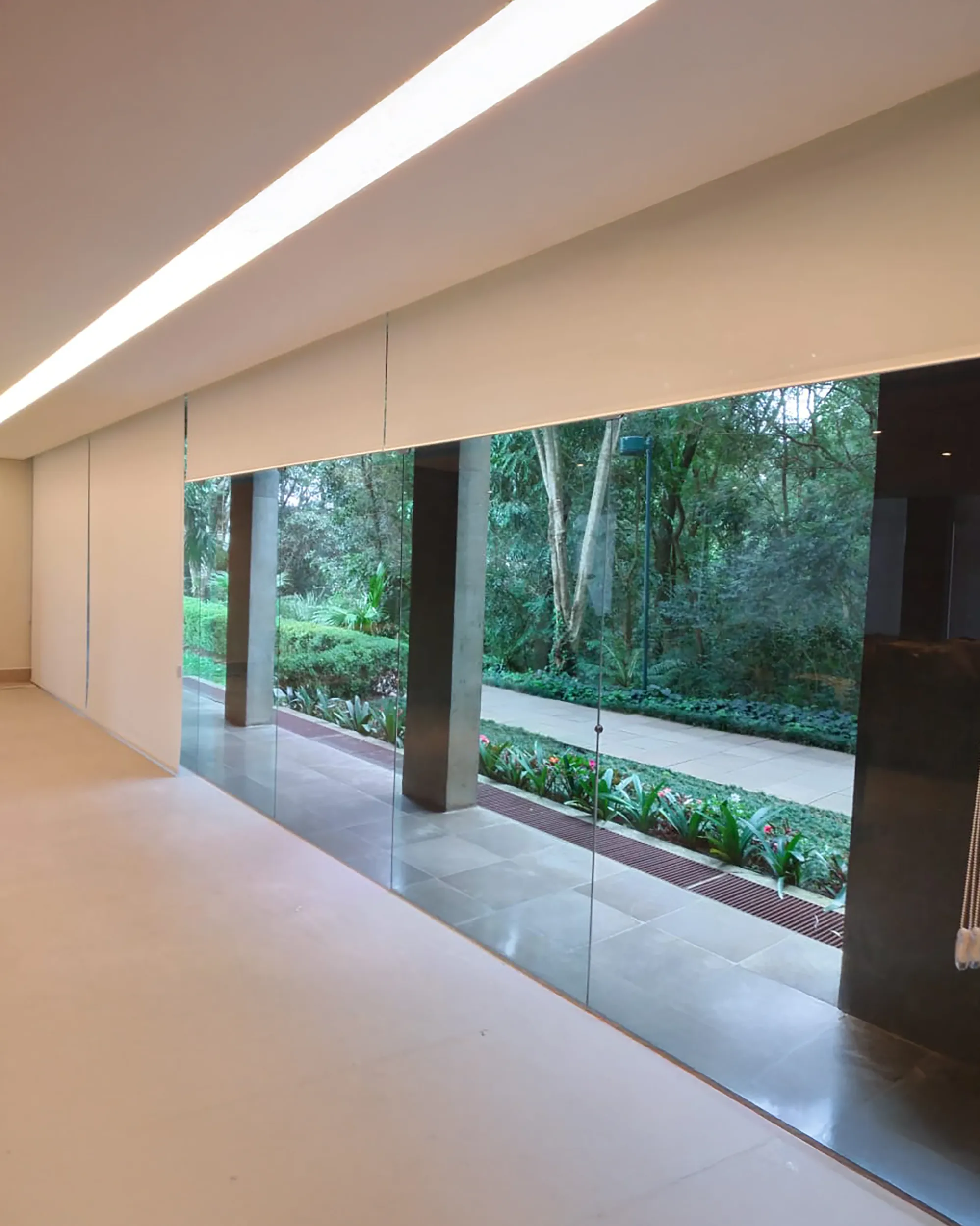 Persiana Rolô Tela Solar 1% branca instalada em grandes vãos de vidro, com vista para o jardim.